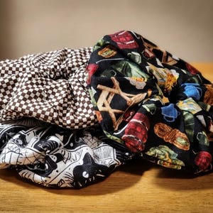 May include: Three fabric hair scrunchies featuring distinct patterns: one with a brown and white checkered print, one with a black background displaying colorful camping gear illustrations, and one with a black and white graphic comic book print.