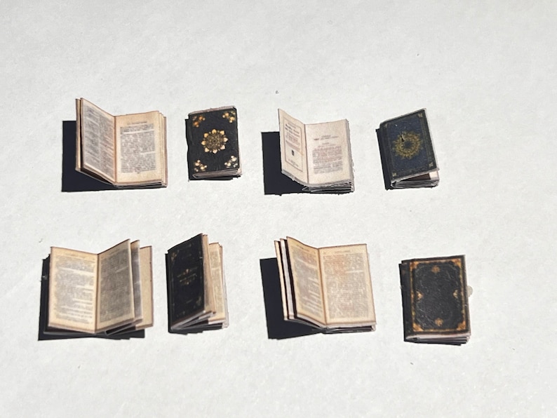 Może przedstawiać: Osiem miniaturowych książek, cztery z otwartymi stronami i cztery zamknięte. Otwarte książki mają strony z tekstem drukowanym czarnym atramentem. Zamknięte książki mają czarne okładki ze złotymi akcentami.
