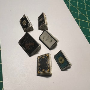 Może przedstawiać: Sześć miniaturowych książek w czarnych, niebieskich i zielonych okładkach. Książki mają złote akcenty i skomplikowane wzory. Książki są ułożone na białym tle.