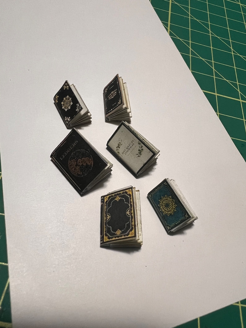 Może przedstawiać: Sześć miniaturowych książek w czarnych, niebieskich i zielonych okładkach. Książki mają złote akcenty i skomplikowane wzory. Książki są ułożone na białym tle.