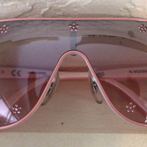 May include: Pink aviator sunglasses with rhinestones and the text "Ray-Ban" and "RJ952058 2241/E 2N" on the frame.