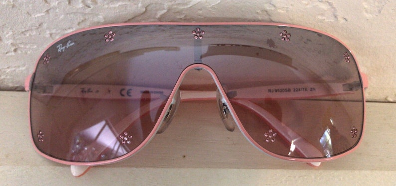 May include: Pink aviator sunglasses with rhinestones and the text "Ray-Ban" and "RJ952058 2241/E 2N" on the frame.