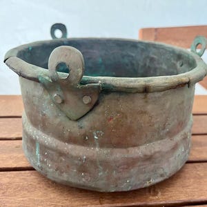 May include: A weathered, antique copper bucket with a rounded body and two small handles. The bucket has a tarnished, aged appearance with hints of green oxidation. The rim is slightly bent, adding to its rustic charm. A decorative piece.