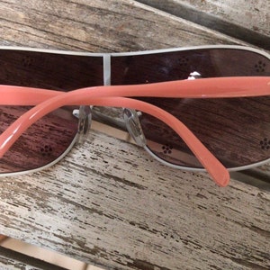 May include: A pair of pink and silver Ray-Ban sunglasses with brown lenses. The sunglasses have a floral design on the arms.