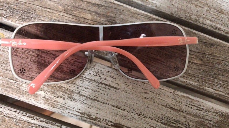 May include: A pair of pink and silver Ray-Ban sunglasses with brown lenses. The sunglasses have a floral design on the arms.