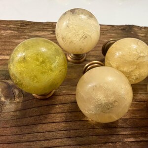 May include: A collection of spherical cabinet knobs featuring translucent, textured stone-like centers in shades of pale yellow and lime green, each mounted on a circular metallic base with a warm, muted bronze finish.