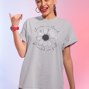 May include: A light gray t-shirt featuring a daisy design with the words "United we stand, divided we fall." The daisy has a black center and white petals with the word "united" written on each petal. The shirt is made of a soft material.