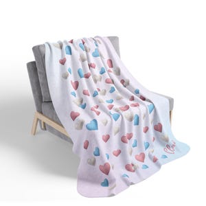 May include: A white blanket draped over a gray chair, adorned with a pattern of pink, blue, and gray hearts. The blanket has a soft, textured appearance, with a light blue gradient at the bottom. The word "Claire" is written in cursive on the blanket.