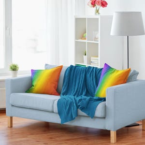 May include: A light blue sofa with two rainbow gradient throw pillows and a blue knitted throw blanket. The sofa has wooden legs and is in a well-lit living room with a lamp and a white bookshelf.