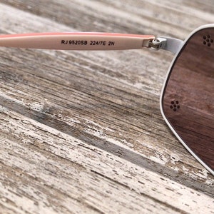 May include: White sunglasses with a pink frame and brown lenses. The sunglasses have the text "RJ 952058 224/7E 2N" printed on the frame.