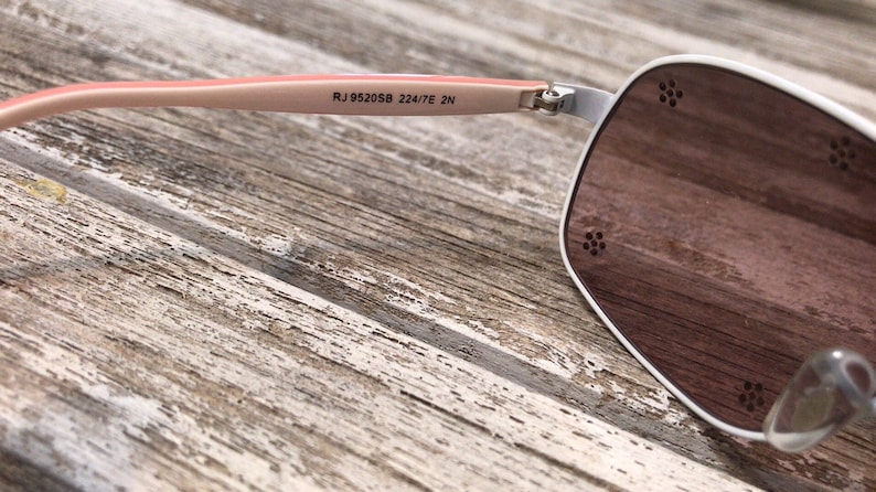 May include: White sunglasses with a pink frame and brown lenses. The sunglasses have the text "RJ 952058 224/7E 2N" printed on the frame.