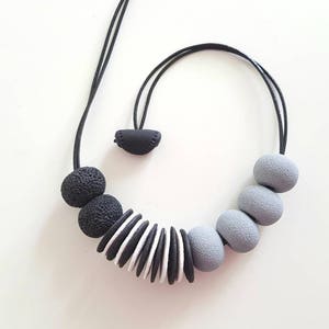 以下が含まれることがあります： 黒とグレーの丸いビーズと平らな丸いビーズを重ねたネックレス。ネックレスはポリマークレイ製で、黒い紐が付いています。