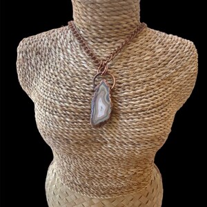 May include: A copper chain necklace with a large agate pendant. The agate is a light brown color with white and gray bands. The pendant is attached to the chain with a copper ring.