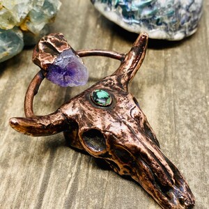 May include: A copper bull skull pendant with a purple amethyst stone and a green turquoise stone. The pendant is on a wooden surface.