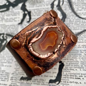 May include: A copper cuff bracelet with a large, natural agate stone set in the center. The stone has a swirling pattern of orange, white, and brown. The bracelet has four copper rivets around the edge.