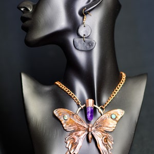 May include: A copper butterfly necklace with an amethyst crystal pendant and turquoise accents. The necklace is displayed on a black mannequin bust.