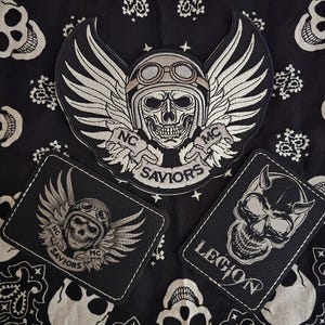 May include: Three black and white patches with skull designs. The largest patch features a skull wearing a helmet with wings and the text "NC SAVIORS MC". The other two patches have similar skull and wing designs, with one reading "NC SAVIORS MC" and the other "LEGION".
