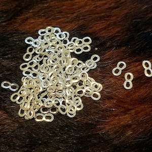 May include: A collection of small, silver-colored infinity connectors scattered on a brown and black textured surface. The connectors are in the shape of the number eight, and are made of a shiny metal. The connectors are likely for jewelry making or crafts.