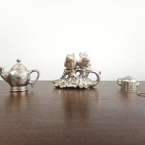 May include: Three silver teapot salt and pepper shakers. One shaker is a traditional teapot design, another is a teapot with a strainer, and the third is a pair of pig salt and pepper shakers on a floral base.