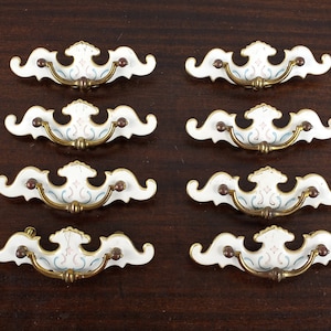 May include: Eight white porcelain cabinet knobs with gold accents and a floral design. The knobs have a curved shape and are designed for use on drawers or cabinets.