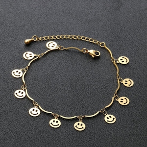 Puede incluir: Un tobillo dorado con un diseño de cadena y múltiples dijes con caritas sonrientes.