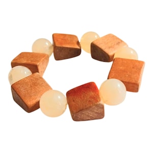 Può includere: Un braccialetto elastico caratterizzato dall'alternanza di perline cubiche in legno marrone e perline rotonde, lisce e traslucide color bianco sporco. Le perline in legno hanno una finitura opaca e testurizzata, mentre quelle rotonde sono lucide.
