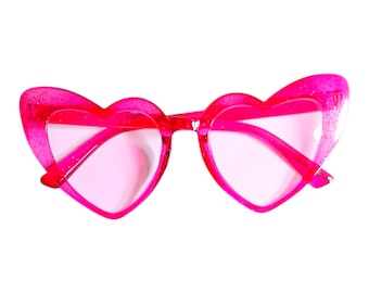 May include: Bright pink heart-shaped sunglasses with a glittery finish. The frames and lenses are a matching shade of pink. The sunglasses are set against a white background.