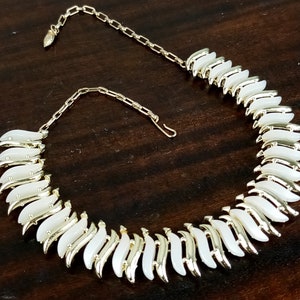 May include: A vintage necklace with a gold-tone chain and white enamel leaf-shaped links. The necklace is displayed on a dark brown wooden surface, showcasing its elegant design and craftsmanship. The clasp is visible.