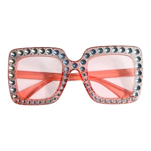 Puede incluir: Gafas de sol cuadradas de gran tamaño con un tinte rosa claro. Las monturas son de color rosa claro y están adornadas con pequeños pedrería plateada. Estas gafas de sol tienen una estética glamurosa y retro.