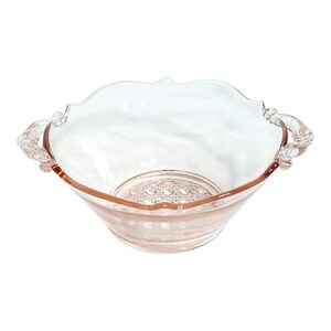 May include: A clear, pink-tinted glass bowl with a scalloped rim and decorative handles. The bowl has a circular, patterned base and a slightly flared shape. The rim and handles have a darker pink hue.