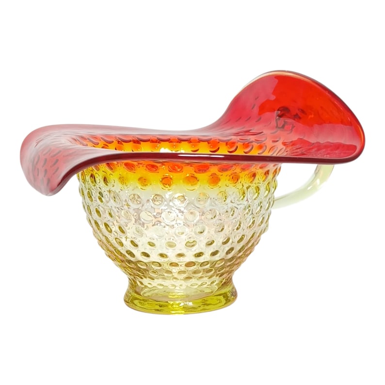 It features a distinctive "Jack-in-the-Pulpit" silhouette with an exaggerated, hand-forged rim and an applied handle. 
The glass exhibits an amberina effect, a color-changing fade from yellow/amber at the base to a vibrant red at the top. 
The body of the piece has a classic hobnail pattern, which consists of raised bumps across the surface. 
Kanawha Glass Company produced these pieces in the mid-20th century, typically in the 1950s to 1970s.