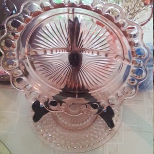 May include: Pink glass serving dish with three compartments and a decorative scalloped edge. The dish is supported by a black stand and sits on a matching base. The dish is likely used for serving appetizers or desserts.