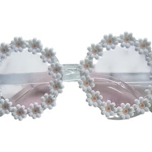 May include: Clear-framed sunglasses with pink lenses, adorned with a ring of white daisy flowers around each lens. The sunglasses have a transparent bridge and temples, creating a whimsical and floral accessory.