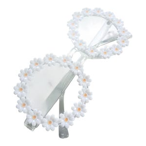 May include: Clear-framed sunglasses adorned with white daisy flowers around the lenses. The sunglasses have a classic shape with a modern floral twist, perfect for adding a touch of whimsy to any outfit.