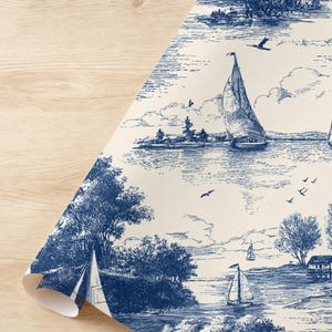 May include: A roll of wallpaper featuring a blue and white toile design. The pattern depicts a serene waterscape with sailboats, trees, and buildings. The paper is partially unrolled, revealing the design against a light wood surface.
