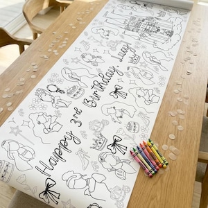 May include: A white coloring roll with black line drawings of princesses, crowns, and carriages. The text "Happy 3rd Birthday, Lucy!" is written on the roll. A bundle of crayons sits on the wooden table next to the roll.