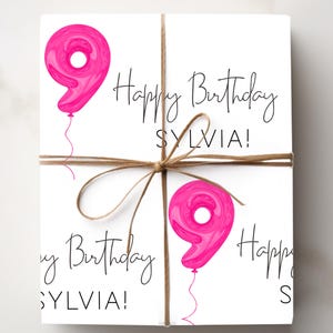 Może przedstawiać: Zapakowany prezent z białym tłem, zawiązany sznurkiem. Prezent jest ozdobiony r&oacute;żowymi balonami z numerem dziewięć i napisem "Happy Birthday SYLVIA!" czarnym pismem.