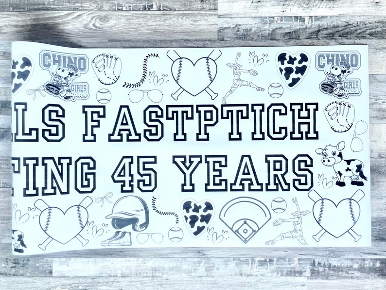 May include: A white banner with black text and graphics celebrating 45 years of Girls Fastpitch. The banner features a baseball diamond, bats, hearts, and cow graphics. The text on the banner reads "Girls Fastpitch Celebrating 45 Years".