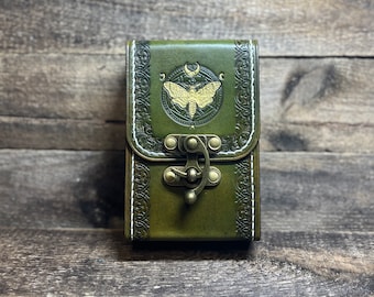 May include: A green leather card holder with a decorative embossed design and a brass clasp. The front features a gold-colored moth and moon design.