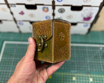 以下が含まれることがあります： 真鍮の留め金が付いた小さな茶色の革箱。箱は質感のある表面で、手で持たれています。