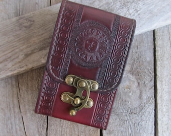 May include: A burgundy leather case with a sun design embossed on the front. The case has a brass clasp and decorative border. The case is rectangular and appears to be a small wallet or container.
