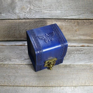 May include: A small, blue leather box with a gold clasp and a raised, embossed design of the Triforce symbol from the Legend of Zelda video game series.