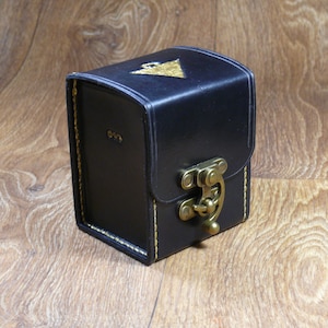 Peut inclure: Une petite boîte en cuir noir avec un fermoir doré et un motif de triangle doré sur le dessus. La boîte a des détails de couture jaune autour des bords.