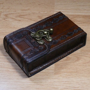 Peut inclure: Un étui en cuir marron avec des motifs floraux complexes et un fermoir en laiton. L'étui est rectangulaire et fermé.