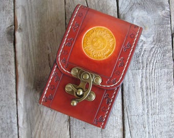 May include: A red leather phone case with a sun design and a brass clasp. The case has a decorative stitched border.
