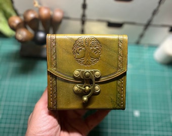 May include: A small, green leather box with a tree design embossed on the front. The box has a brass clasp and a decorative border.