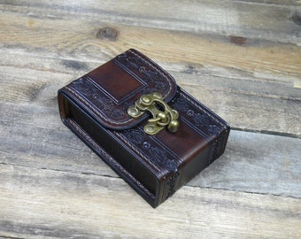 May include: A small, brown leather box with a decorative embossed design and a brass clasp. The box is closed and sitting on a wooden surface.