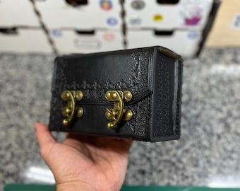 May include: A black leather box with ornate floral detailing and antique brass clasps. The box is rectangular with a hinged lid and a decorative border. The box is held in a hand.