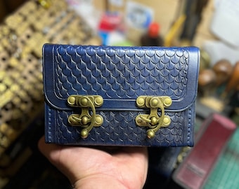 May include: A rectangular dark blue leather box featuring an embossed scale pattern across the exterior surface. The box is secured with two antique brass-colored metal swing latches attached to the front flap, providing a functional closure.