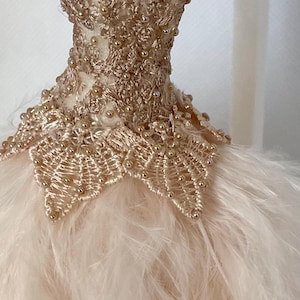Puede incluir: Un vestido en miniatura, probablemente para una muñeca, cuelga de una percha plateada. El corpiño está intrincadamente detallado con bordados y abalorios dorados. La falda está hecha de suaves plumas de color rosa claro, creando una apariencia delicada y elegante.
